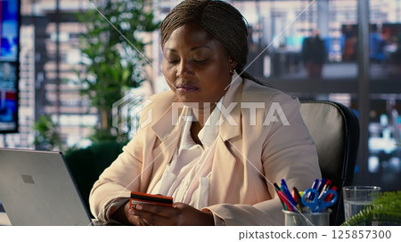 African american CEO entering credit card numbers on her webpage, doing online shopping in office workspace. Director with a success story making an electronic purchase, business success. Camera A. 125857300