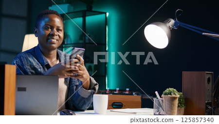African american woman relaxing in apartment, texting friends on cellphone, enjoying leisure time. Smiling remote worker taking break to talk with mates on internet using phone, camera B African american woman relaxing in apartment, texting friends on cellphone, enjoying leisure time. Smiling remote worker taking break to talk with mates on internet using phone, camera B 125857340
