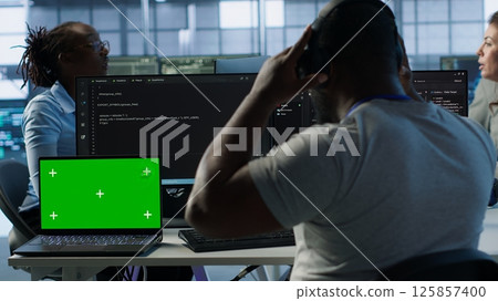 Isolated screen laptop next to programmer coding in server farm while listening music in headphones. Worker safeguarding hardware against unauthorized access with chroma key notebook, camera A 125857400