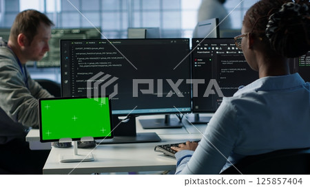 Isolated screen tablet in front of IT support workers coding on computers, upgrading equipment. Engineers at PC desk overseeing server clusters using chroma key device, camera A Isolated screen tablet in front of IT support workers coding on computers, upgrading equipment. Engineers at PC desk overseeing server clusters using chroma key device, camera A 125857404