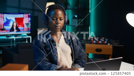 Freelancer doing computer tasks for team project in personal home office while listening music. Remote employee working late at night, typing on laptop keyboard, putting headphones on, camera B 125857405