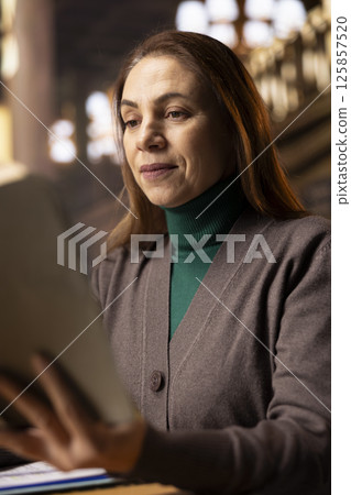 Mature university student preparing her thesis in a campus library study room, reading a book to collect academic information. Ambition and determination to complete a college degree. Mature university student preparing her thesis in a campus library study room, reading a book to collect academic information. Ambition and determination to complete a college degree. 125857520