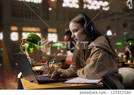 University scholar listening to therapy music on headset to create the perfect ambient for research, working on her diploma paper. Young girl uses focus music to help with collecting information. University scholar listening to therapy music on headset to create the perfect ambient for research, working on her diploma paper. Young girl uses focus music to help with collecting information. 125857580