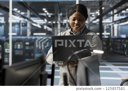Smiling engineer using laptop in data center to oversee hardware delivering computing power for AI training models. Happy worker supervising rackmount servers storing complex machine learning datasets Smiling engineer using laptop in data center to oversee hardware delivering computing power for AI training models. Happy worker supervising rackmount servers storing complex machine learning datasets 125857581