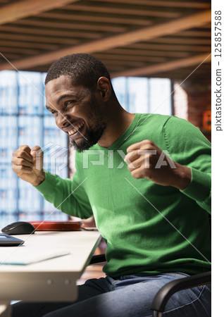 Overjoyed man thrilled by achievement, cheering and gesticulating at home. Happy african american person smiling, celebrating success, showing positive emotions, sitting at apartment desk Overjoyed man thrilled by achievement, cheering and gesticulating at home. Happy african american person smiling, celebrating success, showing positive emotions, sitting at apartment desk 125857588