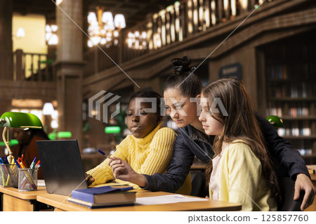 Caucasian tutor guiding two elementary school girls at a library desk, providing private lessons to aid in their academic development with work books and notes. Teacher discussing with scholars. Caucasian tutor guiding two elementary school girls at a library desk, providing private lessons to aid in their academic development with work books and notes. Teacher discussing with scholars. 125857760