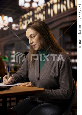 Mature college student completing class notes on printed papers, writing important information in a study room under green lamp. Female adult learner pursuing academic qualifications. 125857761