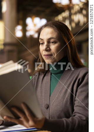 Senior woman reading a book in a study room at a public library, reviewing papers and looking for thesis citations sources. Mature female student determined to graduate college. Intellectual. Senior woman reading a book in a study room at a public library, reviewing papers and looking for thesis citations sources. Mature female student determined to graduate college. Intellectual. 125857871