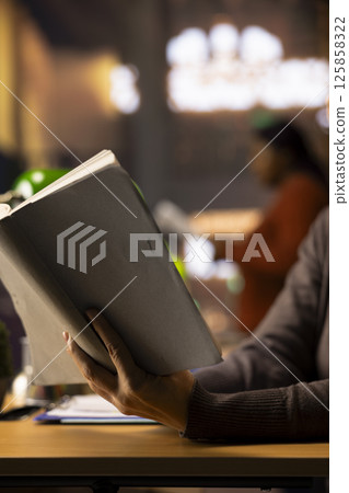 Ambitious female student working on her thesis in a campus library study area, reading a book to collect academic information. Ambition and determination to complete a college degree. Close up. Ambitious female student working on her thesis in a campus library study area, reading a book to collect academic information. Ambition and determination to complete a college degree. Close up. 125858322