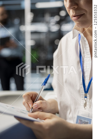 Woman in data center taking note on fixes to be made after running diagnostic scripts on computer. Server room admin writing repairing needed by supercomputers after evaluating performance metrics 125858332