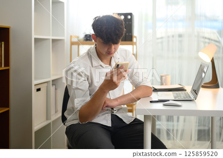 Young man in white using smartphone while working from home with laptop on the desk 125859520