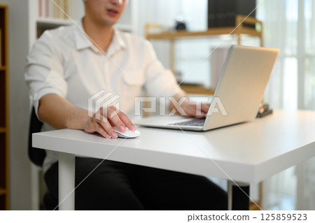Close up of a man using wireless mouse and laptop at white desk working from home 125859523