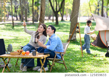 Relaxed family sitting outdoors at a campsite surrounded by trees and fresh greenery 125859633