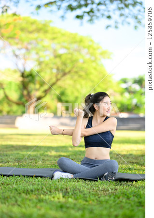 Young woman sitting on a yoga mat and stretching her arm during a workout in the park 125859706