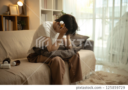 Sad man in a dimly lit living room appearing overwhelmed and anxious with pills in hand 125859729