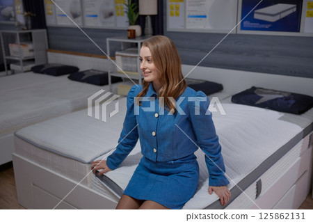 Stylish woman sits on mattress in a contemporary bedding showroom, showcasing comfort and elegance Stylish woman sits on mattress in a contemporary bedding showroom, showcasing comfort and elegance 125862131