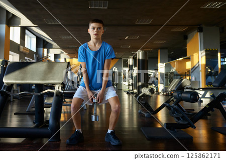 Athletic teenage boy squatting with dumbbells training over gym interior 125862171