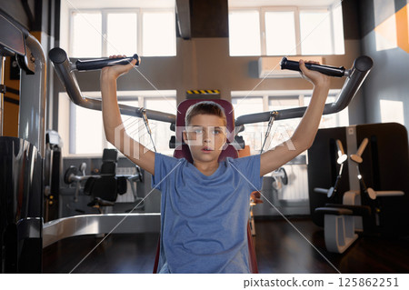 Teenager boy doing sports exercise at machine for chest muscles pumping Teenager boy doing sports exercise at machine for chest muscles pumping 125862251