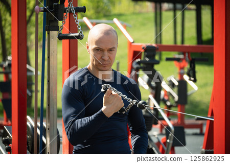 Adult sporty man exercising crossover trainer at public gym park. Outdoors activity. Fitness coach make exercise for arms. 125862925