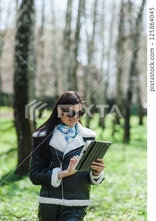 A girl in sunglasses is walking in the park with a tablet. A young woman is working on a tablet in the park in sunny weather. A girl in sunglasses is walking in the park with a tablet. A young woman is working on a tablet in the park in sunny weather. 125864045