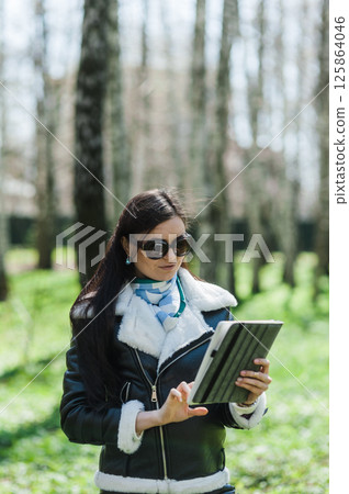 A black-haired woman is flipping through something on a tablet in the park. Young Woman in black glasses and black jacket looking at a tablet in the park. A black-haired woman is flipping through something on a tablet in the park. Young Woman in black glasses and black jacket looking at a tablet in the park. 125864046