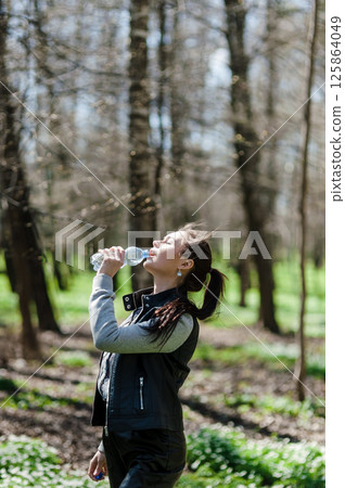 The girl has tilted her head and is drinking mineral water. Beautiful girl in the park drinking water from a bottle. 125864049