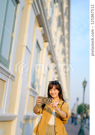 Southeast Asian woman travel concept captures joyful young woman taking selfie outdoors near historic building in warm sunlight expressing happiness and adventure Southeast Asian woman travel concept captures joyful young woman taking selfie outdoors near historic building in warm sunlight expressing happiness and adventure 125867512