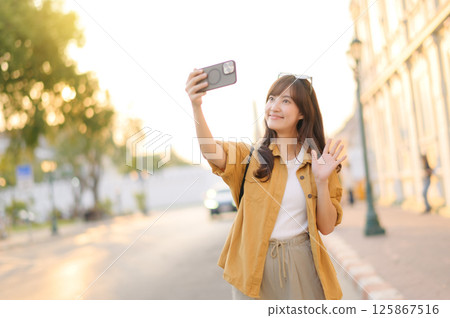 Southeast asian woman enjoys travel concept by taking selfie outdoors with cheerful smile and waving hand in bright urban setting during golden hour 125867516
