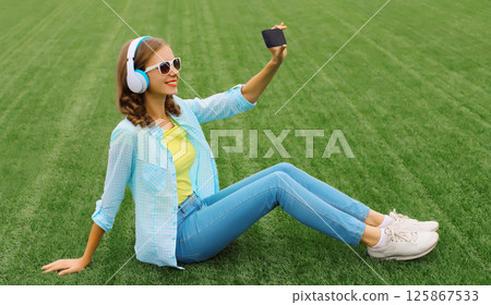 Happy young woman taking selfie with smartphone listening to music lying on grass in summer park 125867533