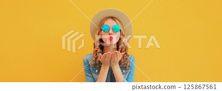 Portrait beautiful stylish young woman blowing kiss in summer hat posing on yellow studio background 125867561