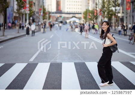Southeast asian woman smiles happily while crossing city street wearing casual black pants and white top with backpack on bright day in urban environment 125867597