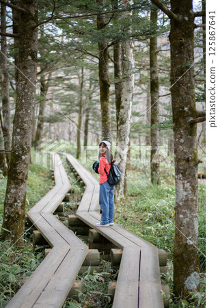 Southeast Asian woman enjoys peaceful walk on wooden path through lush forest wearing casual clothes and backpack with joyful expression on her face Southeast Asian woman enjoys peaceful walk on wooden path through lush forest wearing casual clothes and backpack with joyful expression on her face 125867641