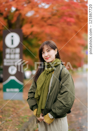 Southeast asian woman wearing green scarf and jacket smiles gently while standing outdoors in autumn with vibrant orange leaves creating warm and peaceful atmosphere 125867720