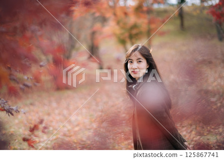 Southeast asian woman smiles gently while standing in serene autumn park surrounded by colorful red and orange leaves creating warm and peaceful atmosphere Southeast asian woman smiles gently while standing in serene autumn park surrounded by colorful red and orange leaves creating warm and peaceful atmosphere 125867741