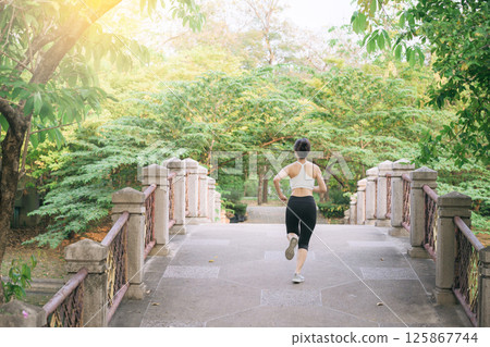 Southeast asian woman jogging in park shows wellness and healthy lifestyle concept with exercise in nature, surrounded by green trees and morning sunlight for peaceful mood 125867744
