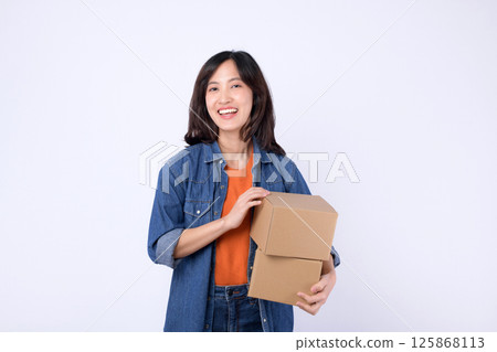 Happy southeast asian woman shopping online holds two cardboard boxes smiling brightly in casual denim jacket and orange shirt against plain white background Happy southeast asian woman shopping online holds two cardboard boxes smiling brightly in casual denim jacket and orange shirt against plain white background 125868113