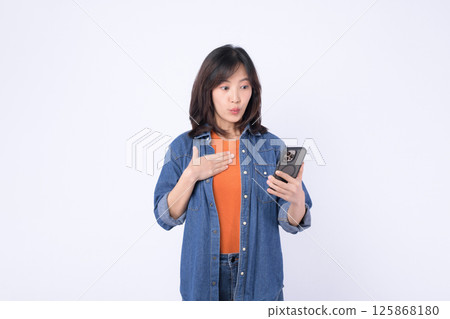 Southeast asian woman uses mobile phone with internet application looking surprised and engaged in casual denim clothing against white background 125868180