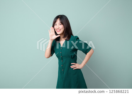 Cheerful southeast asian woman in green dress is advertising with bright smile and hand near her mouth on plain background 125868221