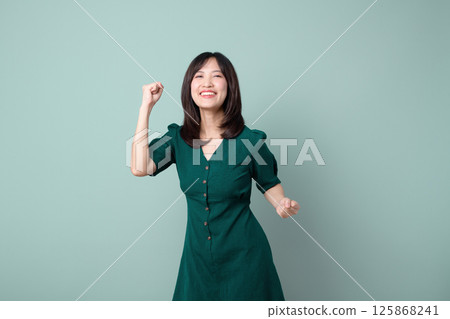 Southeast asian woman in green dress expressing joy and confidence for advertising with bright smile and raised fist on plain background Southeast asian woman in green dress expressing joy and confidence for advertising with bright smile and raised fist on plain background 125868241