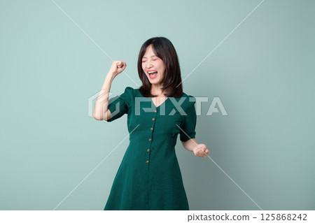 Happy southeast asian woman celebrating with closed eyes and fist raised in joy wearing green dress for advertising concept Happy southeast asian woman celebrating with closed eyes and fist raised in joy wearing green dress for advertising concept 125868242