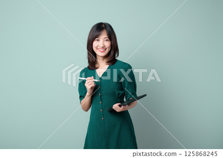 Southeast Asian woman smiling confidently while holding digital tablet and stylus for advertising or creative work in green dress 125868245