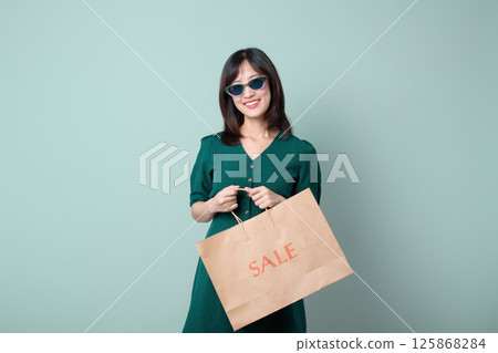 Southeast asian woman smiles happily wearing sunglasses and holding shopping bag with sale text while using mobile internet application for shopping convenience 125868284