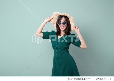 Southeast asian woman wearing sunglasses and straw hat smiles brightly in this advertising style portrait with green dress and light background 125868295