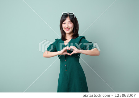 Southeast asian woman in green dress smiling happily while making heart shape with her hands for advertising on plain background 125868307