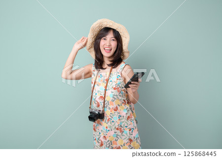 Southeast asian woman joyfully uses mobile phone to explore internet application while wearing floral dress and straw hat with camera hanging around her neck 125868445
