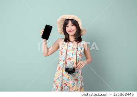 Southeast asian woman wearing floral dress and straw hat happily shows her phone screen while holding camera around her neck, enjoying online shopping and travel moments Southeast asian woman wearing floral dress and straw hat happily shows her phone screen while holding camera around her neck, enjoying online shopping and travel moments 125868446