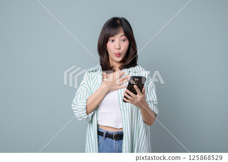 Southeast asian woman uses mobile phone with internet application looking surprised and excited while standing against plain background in casual clothing Southeast asian woman uses mobile phone with internet application looking surprised and excited while standing against plain background in casual clothing 125868529