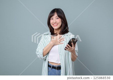 Southeast asian woman smiles happily while holding mobile phone using internet application showing positive emotion and casual style in front of plain background 125868530