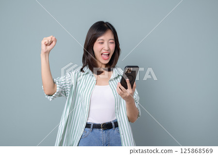 Southeast asian woman excitedly uses mobile phone to access internet application, celebrating joyful moment with raised fist and happy expression in casual outfit Southeast asian woman excitedly uses mobile phone to access internet application, celebrating joyful moment with raised fist and happy expression in casual outfit 125868569
