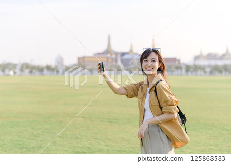 Southeast asian woman smiles happily while travel outdoors in green field with cityscape background capturing travel concept with casual outfit and backpack 125868583